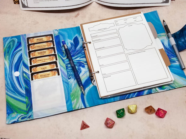 Open binder showing the inside of the front cover. Six card slots are inside. the opposing page shows page one of the character sheet, dividers and a pen