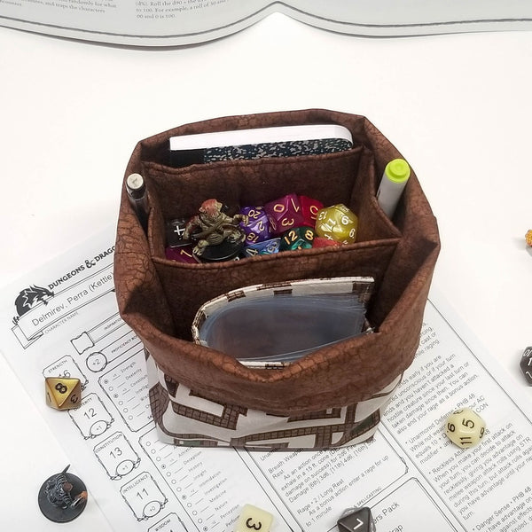 brown and cream dungeon map print dice bag. Top folded down to expose brown lining and pockets. Notebook, spellbook, dice and pencils in the pockets.