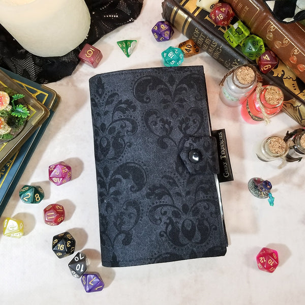 Tarot sized spellbook/card album lies flat on a table surrounded by dice, vintage books, minis and potions